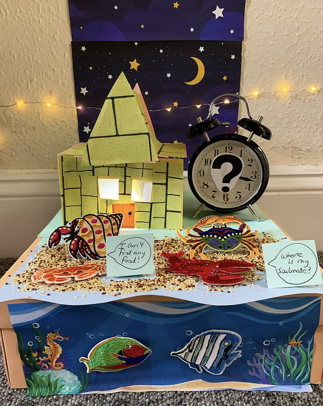 Two cartoon sea creatures standing in front of a cardboard house (lit from inside) and a clock (with part of its face covered by a question mark). Behind this arrangement is a poster featuring a night sky. The left-hand creature is saying 'I can't find any food'. The right-hand creature is saying 'Where is my soulmate?'. 