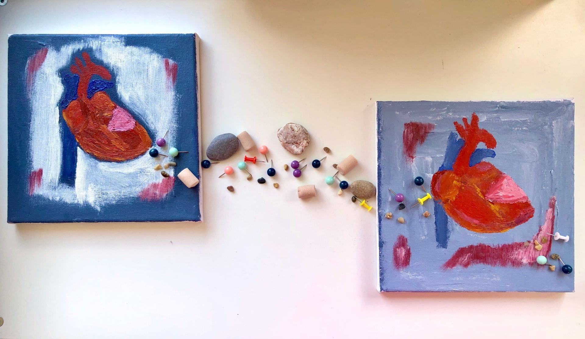 Two square paintings of hearts. Between them is an assortment of stones, earplugs and drawing pins.
