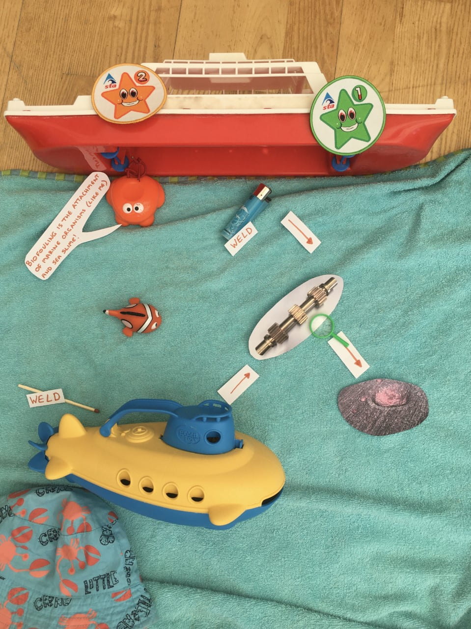 A toy boat with a towel beneath it. On top of the towel, which is intended to look like the sea, are: a lighter (labelled 'weld'); a toy frog (which is saying 'biofouling is the attachment of marine organisms (like me) and sea slime!); some toy fish; a metal gear; a toy submarine; a shell. Next to the submarine is a match (labelled 'weld').