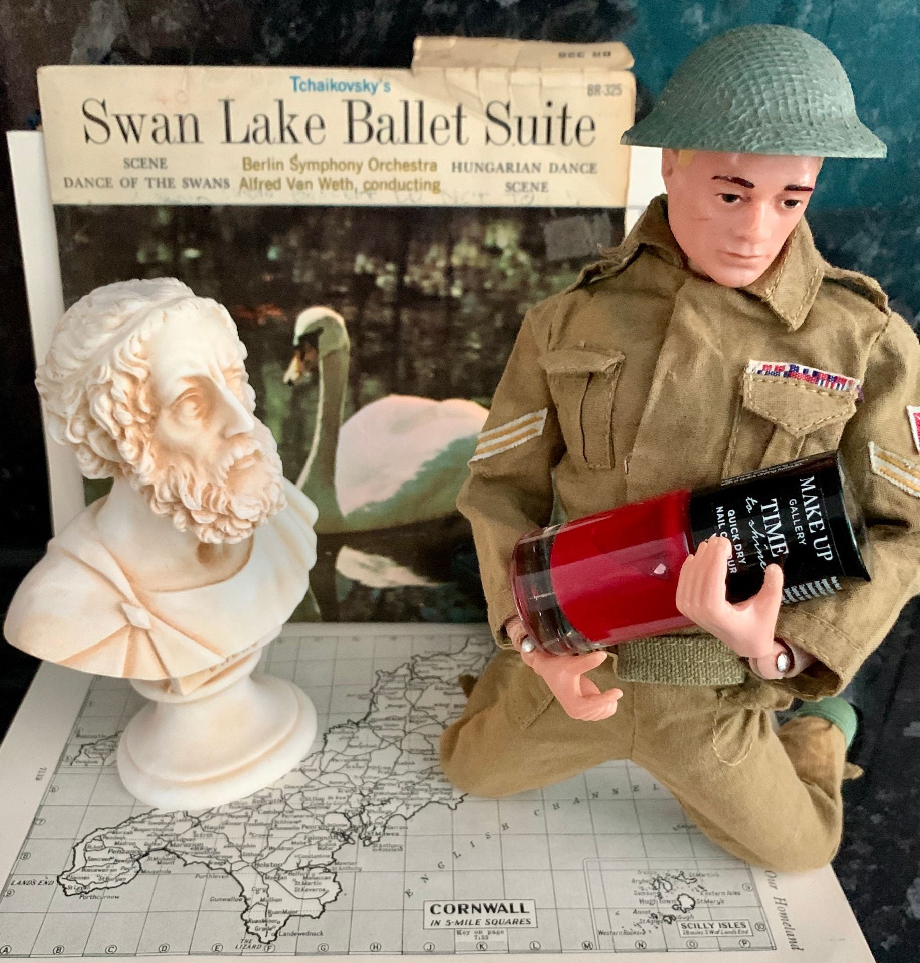 A classical bust and an 'Action Man' toy (holding nail varnish) on top of a map of Cornwall. Behind them is a the sleeve of a vinyl record: Tchaikovsky's Swan Lake Ballet Suite.