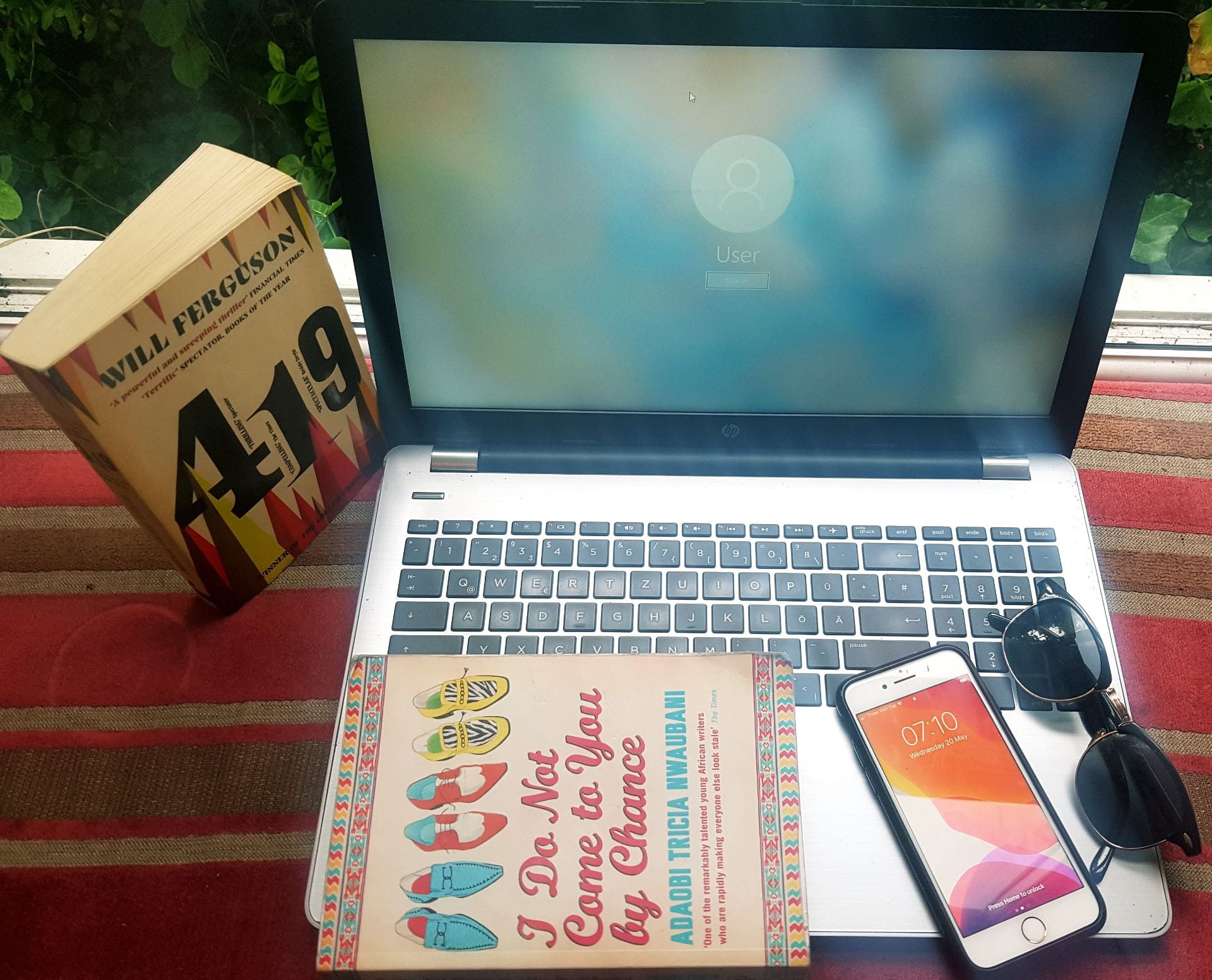A MacBook laptop. On top of it is a novel ('I Do Not Come to You by Chance' by Adaobi Tricia Nwaubani), an iPhone and a pair of sunglasses.. To the left of the MacBook is another novel: '419' by Will Ferguson.