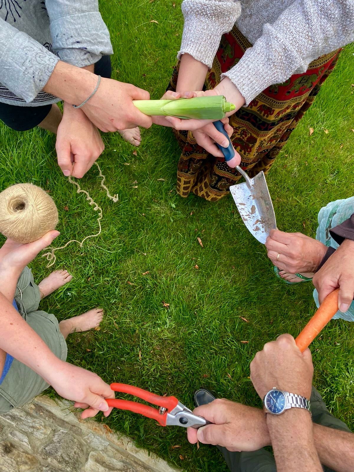 Four people standing in a circle, photographed from above. Their arms are crossed in an 'Auld Lang Syne' style. However, instead of holding each other's hands, they hold objects. The objects are (clockwise from top): a leek; a trowel; a carrot; pruning shears; yarn.