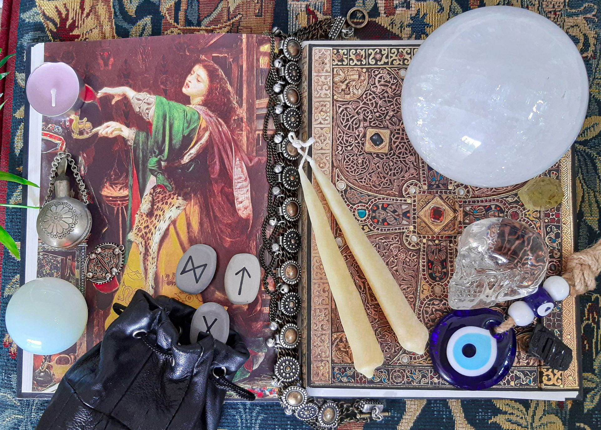 An antique book open on an illustration of a woman. On top of the book are a selection of objects: candles, an open bag of runes; a large, opaque crystal sphere; a transparent crystal skull.